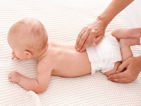 bebê deitado de bruços e uma mão segundo sua perna e outra massageando sua lombar