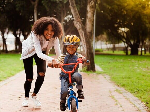 Foto de um menino sorrindo, em uma bicicleta sem rodinhas e ao lado uma mulher adulta também sorrindo segurando em um dos lados do guidão da bicicleta.