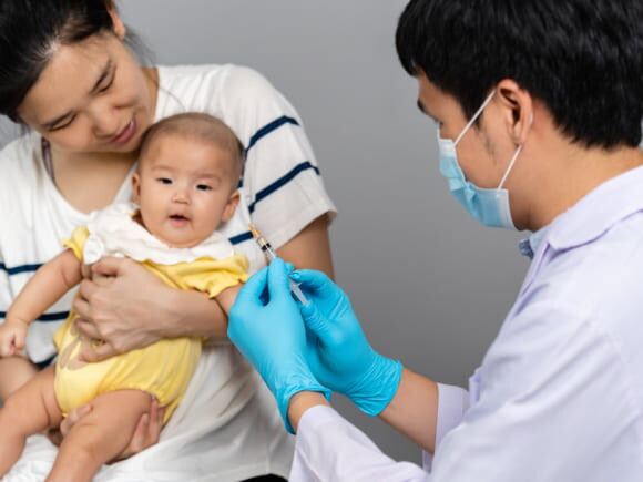 imagem de um bebê no colo de uma mulher adulta e um homem segurando uma seringa, vestindo roupas de profissionais da saúde na cor branca