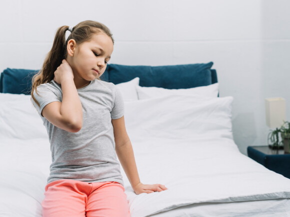 imagem de uma menina sentada na beira de uma cama, com uma das mãos no pescoço e com olhos fechados