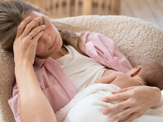 Imagem de uma mãe amamentando seu bebê com a mão na testa e semblante de cansada
