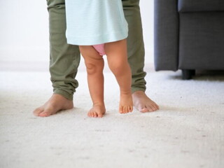 pai, usando calça verde, ajudando o filho a dar os primeiros passos 
