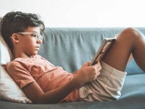 Criança de 7 anos lendo um livro deitada em um sofá, apoiada em uma almofada.