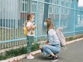 Uma mãe está agachada em frente a sua filha, perto de um portão. A menina está de mochila, olhando para a mãe e segurando um urso de pelúcia.