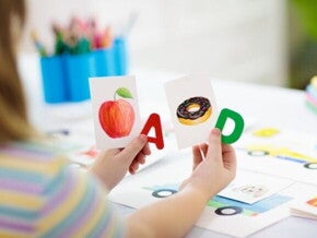 Mãos de criança seguram uma carta A vermelha ao lado de um cartão com uma maçã, e uma carta D, verde, ao lado de um cartão com um donut.