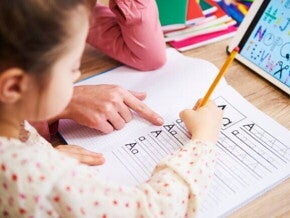 Uma mão adulta aponta para um caderno quadriculado onde uma criança está praticando a escrita das letras "A" e "a" com um lápis.