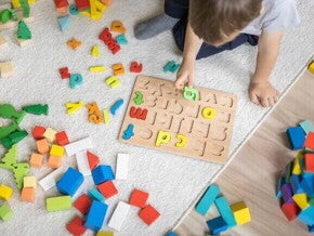 Em um piso de madeira com um tapete cinza, está sentado um menino,vestindo uma camiseta branca. Ele está brincando com um tabuleiro de encaixar letras, enquanto segurando uma delas. Há letras e outras peças coloridas espalhadas ao seu redor.