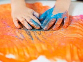Imagem das mãos de uma criança, sentindo a textura de um glitter e de tintas. Ao fundo, um cartaz branco com algumas pinturas na cor laranja, rosa e glitter azul.