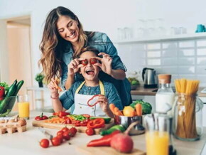 Uma mãe e sua criança estão na cozinha, com diversos alimentos sobre uma bancada. Ela segura dois tomates pequenos na altura dos olhos da criança e ambas estão sorrindo