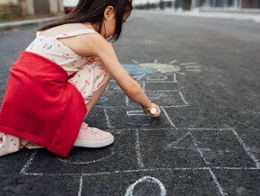 Uma menina de vestido e tênis está agachada na rua desenhando uma amarelinha no chão. A foto faz alusão ao tema de brincadeiras de rua