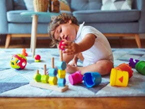 Um bebê está sentado no tapete de uma sala de estar mexendo com brinquedos de empilhar e montar, que é uma das brincadeiras que estimulam o desenvolvimento infantil