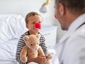 Um menino sorridente com um nariz de palhaço vermelho e segurando um ursinho de pelúcia está sentado em uma cama de hospital, olhando para um médico de costas para o observador. O médico também usa um nariz de palhaço vermelho.