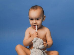 Bebê sentado com uma escova de dentes na boca, segurando um urso de pelúcia Bebê sentado com uma escova de dentes na boca, segurando um urso de pelúcia