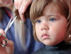 Uma menina recebendo um corte de cabelo. Ao seu lado, a mão de uma mulher segura uma mecha do seu cabelo, enquanto outra mão segura uma tesoura pequena