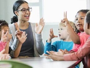 Imagem de uma tutora interagindo com crianças ao redor de uma mesa. Brincadeiras entre professores e crianças ajudam na adaptação na creche dos pequenos