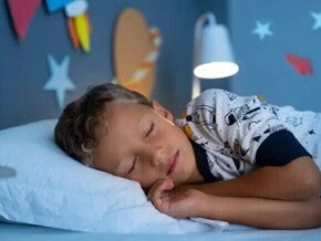 Um menino, vestido de pijama estampado, está deitado na cama, de olhos fechados. O abajur atrás dele está aceso, e nas paredes do quarto há decorações de planetas. 