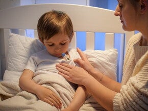 Criança deitada na cama enquanto uma mulher afere sua temperatura com um termômetro