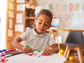 Uma criança de 4 anos está em uma sala de aula, sentado à mesa, e usa uma caneta hidrocor para desenhar