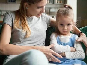 Mãe e filha sentadas no sofá da sala, mãe abraça a criança e conversa, criança parece estressada com os braços cruzados e olhar distante
