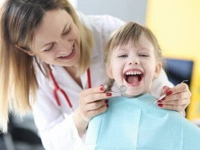 Criança sentada com a boca aberta, ao lado de uma dentista que está em pé, sorrindo e segurando aparelhos odontológicos. Criança sentada com a boca aberta, ao lado de uma dentista que está em pé, sorrindo e segurando aparelhos odontológicos.