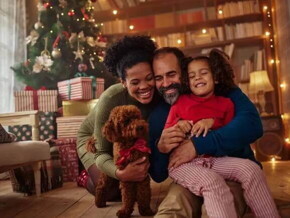 Homem e mulher abraçados com a filha e um cachorrinho no chão, na sala de casa em frente a uma árvore de natal