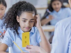 Imagem de uma criança com dislexia olhando para uma ficha amarela com letras B maiúscula e minúscula. Em uma mão ela segura um lápis e a outra está sobre a cabeça em uma postura de dúvida. 