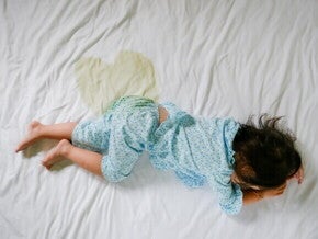 Criança deitada de bruços em uma cama com lençol branco, com uma mancha de xixi visível ao lado, vestindo pijama azul claro com estampa.