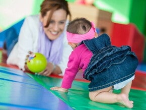 Um bebê engatinhando em direção a uma mulher sorridente, que observa o bebê com uma bola verde na mão. Um bebê engatinhando em direção a uma mulher sorridente, que observa o bebê com uma bola verde na mão.