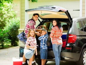 Família posando e sorrindo para a câmera, enquanto organiza malas dentro de um carro preto para sair de viagem