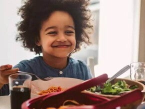 ferro na alimentacao infantil criança sentada a mesa no horário de almoço, sorrindo enquanto segura o talher na mão. Em sua frente estão os pratos com alimentos, incluindo uma salada verde, e há a presença de um adulto do outro lado da mesa.
