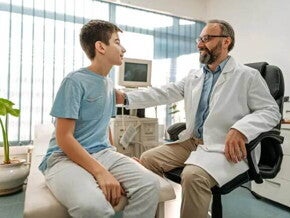 Imagem de um ambiente hospitalar em que um médico tranquiliza um paciente menino, ao colocar a mão no ombro dele. O médico está de jaleco branco, óculos e tem barba e o menino está vestindo uma camiseta azul