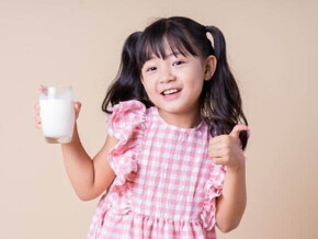 Menina sorrindo, vestindo um vestido xadrez rosa e branco, segurando um copo de leite e fazendo um joinha