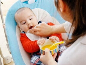 Bebê sentado em uma cadeirinha, com as mãos em um potinho de comida, sendo limpo por um adulto utilizando uma fralda de boca.