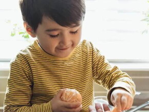 Um menino, vestido com camisa listrada, segura uma tangerina. Entrando no quadro há uma mão de adulto dando frutas para a criança.