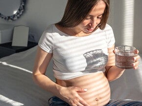 Gestante sentada na cama com uma mão sobre a barriga segurando um copo d’água Gestante sentada na cama com uma mão sobre a barriga segurando um copo d’água