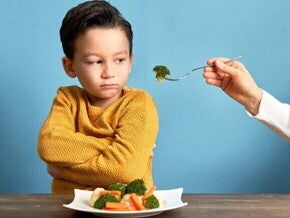 Uma mão estendendo um garfo com brócolis para um menino, que olha de forma irritada para o alimento.