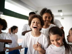 Imagem de um ambiente escolar com várias crianças vestidas de uniforme. O destaque está para um menino gargalhando, bem no centro da foto, de mãos dadas com uma menina