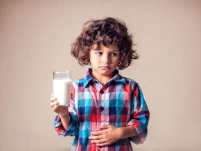 Imagem de uma criança desconfortável, com um copo de leite em uma mão e outra sobre a barriga em uma postura que remete aos efeitos da intolerância à lactose.