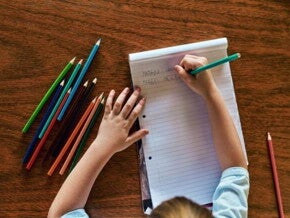 Um menino está escrevendo em um bloco de folhas alguns nomes, como “Fabio”. Na mesa estão espalhados lápis de cor de diversas cores. A foto está ligada ao tema Troca de letras na infância.