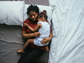 Foto de uma mãe solo dormindo com a filha no colo em sua cama. As duas estão no meio da cama que está com as laterais vazias