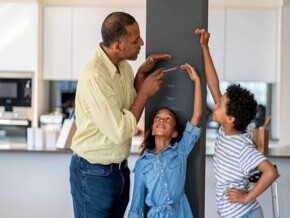 Em uma cozinha, está um homem, uma menina e um menino. A menina está encostada em uma parede, enquanto o homem e o menino marcam na parede qual é a altura dela.