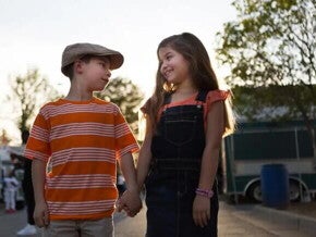  Imagem de um menino e uma menina de mãos dadas se olhando. O menino usa uma boina e uma camisa listrada laranja e branco e a menina está de cabelo solto, usando um macacão escuro e sorrindo