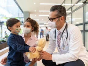 Neuropediatra e enfermeira se aproximam de uma criança, que segura um urso de pelúcia. 
