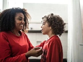 Mulher sorrindo com seu filho, ambos usando um pijama vermelho e conversando. Ao fundo, um quarto.