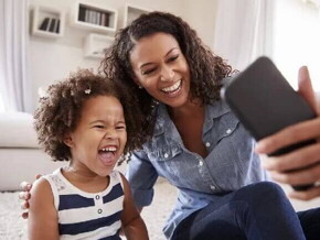 Sentadas no chão, mãe e filha sorriem ao tirar uma foto com o celular. As duas apresentam uma personalidade parecida.