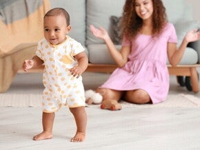 Um bebê sorrindo e em pé no chão, olhando para a frente, com uma mulher sentada no chão ao fundo, sorrindo.