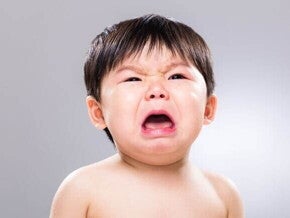 Bebê chorando com expressão intensa de angústia, boca aberta e olhos fechados parcialmente, em um fundo neutro cinza.