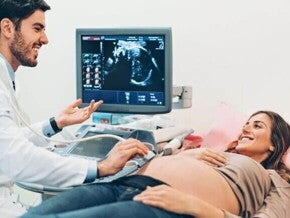 Mulher grávida deitada em uma maca durante exame de ultrassom, enquanto médico realiza o procedimento e conversa com a paciente; ao fundo, monitor exibe a imagem do bebê.