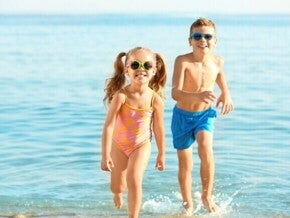 Duas crianças sorrindo e correndo na praia, usando óculos de sol e roupa de banho.