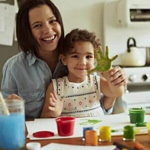 Sentadas à mesa estão uma menina e uma mulher. Espalhadas pela superfície temos tintas de diversas cores e papéis. A mão da criança está toda pintada de verde e as duas estão alegres. A imagem remete a brincadeiras para fazer com crianças mais crescidinhas